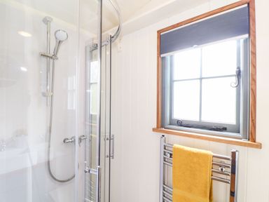 A bathroom with a glass shower enclosure a chrome shower head and a window with a blind and towel rail with a yellow towel at Godrevy Shepherds Hut in Angarrack near Hayle