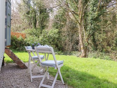 Two white folding chairs with cushions and a small table with flowers outside on gravel beside a grassy area and trees at Godrevy Shepherds Hut in Angarrack near Hayle