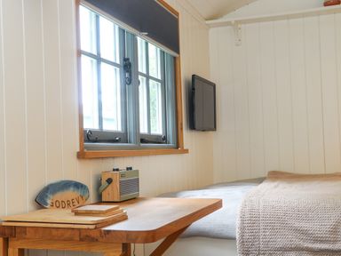A small bedroom with a window a bed a wooden table and a wall-mounted television at Godrevy Shepherds Hut Angarrack near Hayle