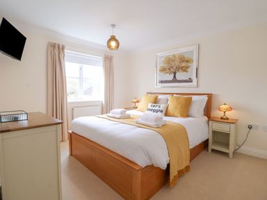 A bedroom with a wooden bed with white and yellow bedding bedside tables with lamps a window and a wall-mounted TV at 23 Church Farm Lane in Halesworth