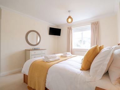 A bedroom with a bed made with white sheets and yellow pillows and throw with a window with curtains and a chest of drawers with a round mirror above at 23 Church Farm Lane in Halesworth