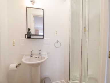 A bathroom with a pedestal sink, mirror with shelf, soap dispenser, toilet paper holder, towel ring, and shower enclosure at 23 Church Farm Lane in Halesworth