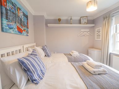A bedroom with two beds white linens striped pillows a wall shelf with decorations hangers on hooks and a window with curtains at 23 Church Farm Lane Halesworth