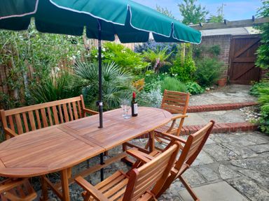 A garden patio with a wooden table and chairs under a green umbrella and plants near a wooden gate at 23 Church Farm Lane in Halesworth