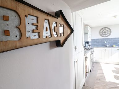A kitchen with white cabinets blue tiled backsplash a large wall clock and a wooden arrow sign saying beach at Ty'n-Y-Ffynnon Cottage in Barmouth