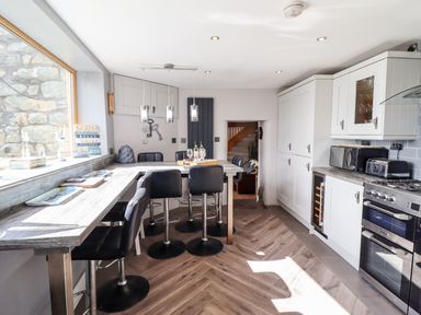 A kitchen with bar stools around a wooden counter and white cabinets at Ty'n-Y-Ffynnon Cottage in Barmouth