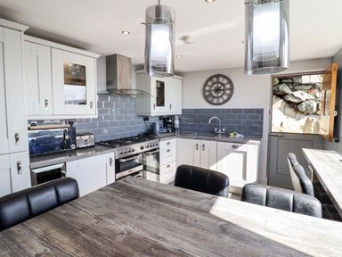 A kitchen with white cabinets a wooden dining table black chairs and a gray tiled backsplash at Ty'n-Y-Ffynnon Cottage in Barmouth