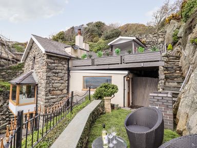 An outdoor seating area with a round glass table and two chairs next to a stone and plaster house with a balcony and stone stairs at Ty'n-Y-Ffynnon Cottage in Barmouth