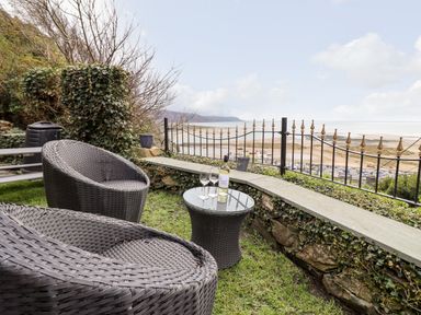 An outdoor seating area with wicker chairs and table overlooking a beach and ocean at Ty'n-Y-Ffynnon Cottage in Barmouth