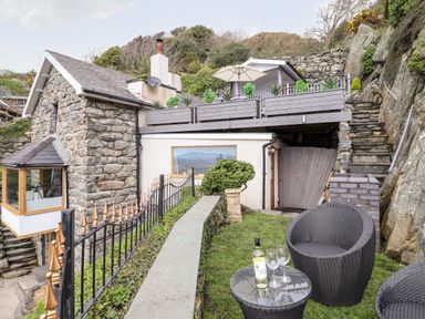 An outdoor seating area with two wicker chairs and a table with a bottle and glasses by a stone house with a balcony and garden at Ty'n-Y-Ffynnon Cottage in Barmouth