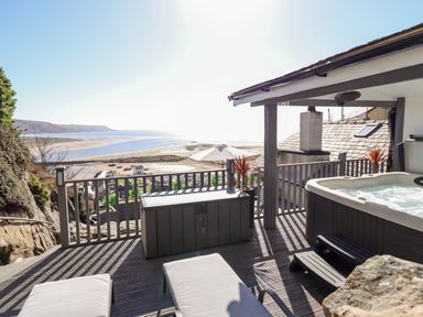 A deck with a hot tub and lounge chairs overlooking a beach and sea at Ty'n-Y-Ffynnon Cottage in Barmouth