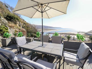 An outdoor seating area with a table umbrella and chairs overlooking a beach and hills at Ty'n-Y-Ffynnon Cottage in Barmouth
