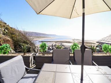 An outdoor patio with chairs and a table under an umbrella overlooking a coastal landscape with hills and water at Ty'n-Y-Ffynnon Cottage in Barmouth