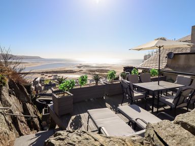 An outdoor terrace with lounge chairs table and umbrella overlooking a beach and ocean at Ty'n-Y-Ffynnon Cottage in Barmouth