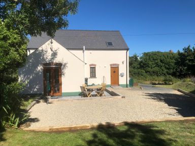 A gravel patio with wooden table and chairs outside a cream house with wooden doors at Ty Rhosyn in Penycwm near Solva