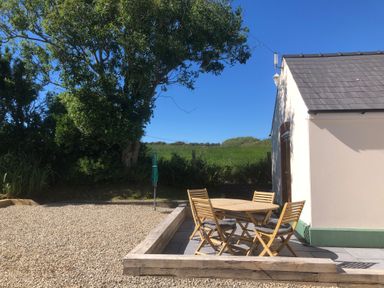 An outdoor patio area with a round wooden table and four chairs next to a small cream building with a gray roof and surrounded by gravel and greenery at Ty Rhosyn Penycwm near Solva