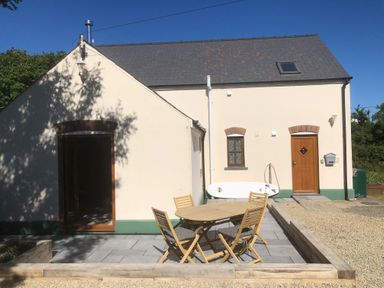 An outdoor patio area with a round wooden table and four chairs outside a cream house with a wooden door and window at Ty Rhosyn in Penycwm near Solva