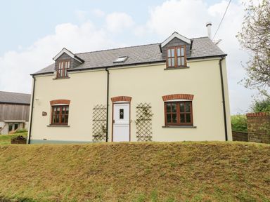 A cream colored two story house with brown framed windows and white door on a sloped grassy hill at Ty Rhosyn in Penycwm near Solva