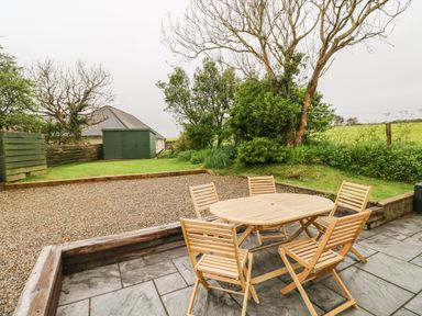 An outdoor patio with a wooden table and chairs next to a garden with trees and a green shed at Ty Rhosyn in Penycwm near Solva