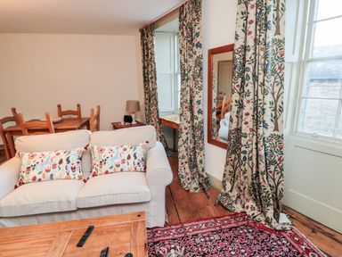 A living room with a sofa, wooden dining table with chairs, patterned curtains, a wooden coffee table, and a large mirror at Upper Oakwood in Alnwick