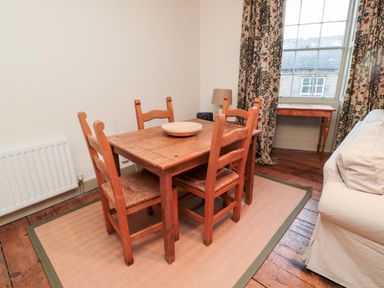 A dining area with a wooden table and four chairs on a rug near a window with floral curtains at Upper Oakwood in Alnwick