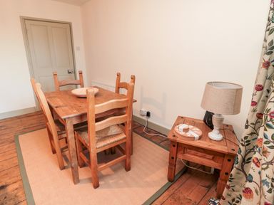 A dining area with a wooden table, four chairs, a side table with a lamp, and floral curtains at Upper Oakwood in Alnwick