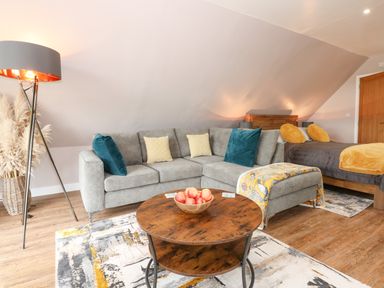 A living area with a grey sectional sofa and a wooden round coffee table with apples a bed with grey bedding and yellow pillows in the background at Strawberry Fields Annex in Emneth