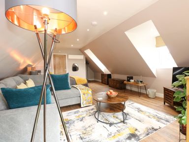 A living room with a gray sofa with teal and cream pillows a wooden coffee table with a bowl of apples a patterned rug slanted ceiling skylights wooden floor and a floor lamp at Strawberry Fields Annex in Emneth