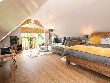 A bedroom with a wooden bed, gray sofa, coffee table, floor lamp, TV on stand, and large triangular window at Strawberry Fields Annex in Emneth