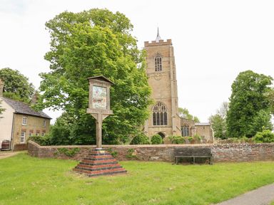 A church tower behind a brick wall with a wooden sign and a bench on grass in Emneth