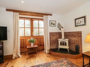 A room with a wooden floor a small stove in a brick corner a table with flowers and wooden doors at Oak Barn in Kentisbeare