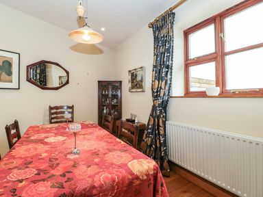 A dining room with a table covered with a floral tablecloth surrounded by chairs and a window with curtains at Oak Barn in Kentisbeare