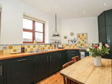 A kitchen with wooden countertops green cabinets a wooden table with flowers and yellow tile backsplash at Oak Barn Kentisbeare