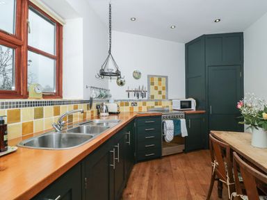 A kitchen with a wooden countertop double sink dark cabinets stove microwave and dining table with chairs and flowers at Oak Barn in Kentisbeare