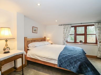 A bedroom with a wooden bed blue quilt curtains and a side table with a lamp at Oak Barn in Kentisbeare