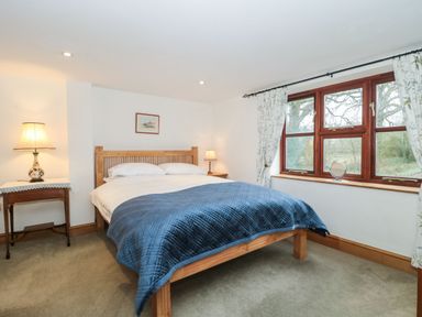 A bedroom with a wooden bed with white pillows and a blue blanket beside a window with floral curtains at Oak Barn in Kentisbeare