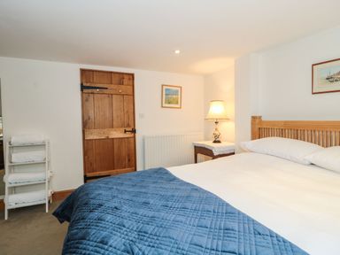 A bedroom with a wooden bed with white pillows and a blue quilt a wooden door a side table with a lamp a towel rack and framed pictures on the wall at Oak Barn in Kentisbeare