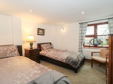 A bedroom with two single beds a wooden nightstand with a lamp a window with curtains a chair and a chest of drawers at Oak Barn Kentisbeare