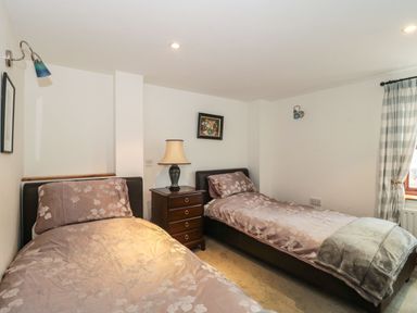 A bedroom with two single beds separated by a wooden nightstand with a lamp and a window with checkered curtains at Oak Barn Kentisbeare