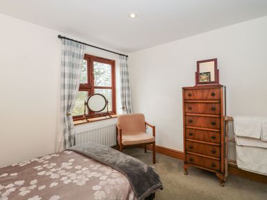 A bedroom with a bed a wooden chair a chest of drawers and a window with checkered curtains at Oak Barn in Kentisbeare