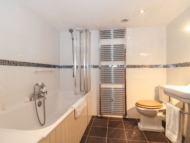 A bathroom with a bathtub and shower, toilet with wooden seat, sink, towel rack, and tiled walls and floor at Oak Barn in Kentisbeare