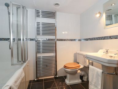 A bathroom with a shower, towel radiator, toilet with wooden seat, and a sink with a mirror at Oak Barn in Kentisbeare