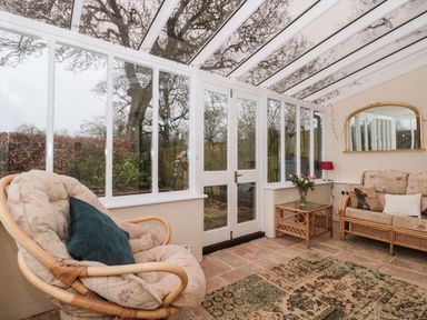 A conservatory with glass walls and ceiling with rattan armchairs a sofa a side table with flowers and a wall mirror at Oak Barn in Kentisbeare