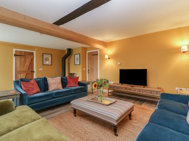 A living room with blue and green sofas a striped ottoman a wooden tv stand with a tv and a vase of flowers on the ottoman at Byways in Madresfield near Malvern