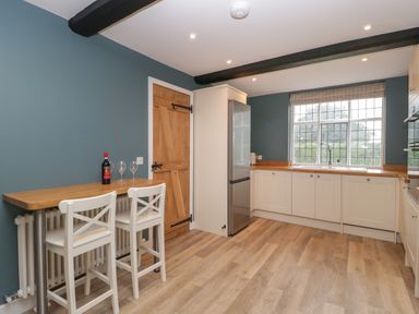 A kitchen with white cabinets wooden countertops a large window a wooden door a refrigerator two white chairs and a wooden breakfast bar at Byways in Madresfield near Malvern