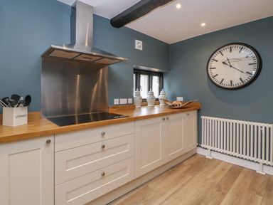 A kitchen with white cabinets a wooden countertop a stainless steel stove and hood utensils in a container and a wall clock at Byways in Madresfield near Malvern