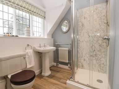 A bathroom with a toilet with dark seat cover a pedestal sink a shower with marble walls a window with a patterned blind and a round mirror on the wall at Byways Madresfield near Malvern