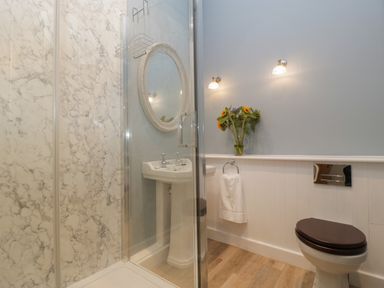 A bathroom with a shower enclosure with marble walls a pedestal sink with a round mirror a towel ring with a white towel a toilet with a dark seat and a vase with flowers at Byways Madresfield near Malvern