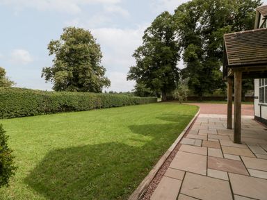 A garden with green grass and large trees beside a paved patio at Byways in Madresfield near Malvern