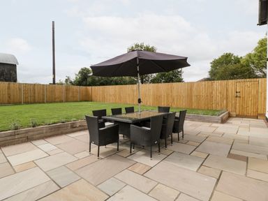 An outdoor patio with a glass table surrounded by eight wicker chairs and a large black umbrella by a lawn and wooden fence at Byways in Madresfield near Malvern